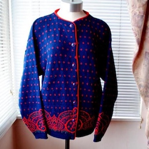 May include: A navy blue cardigan sweater with red diamond patterns and trim. The sweater has a red paisley design at the bottom and red buttons down the front. The sweater is displayed on a mannequin.