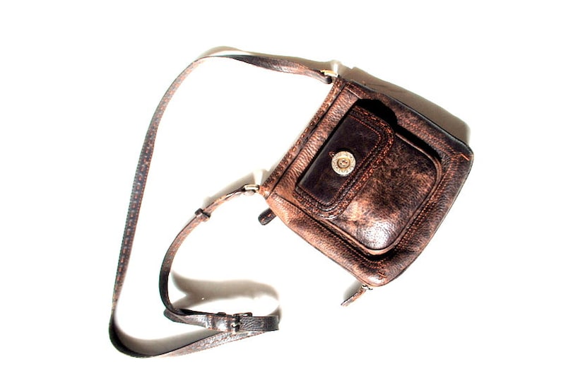 May include: A brown leather crossbody bag with a flap closure and a single pocket on the front. The bag has a worn, distressed look and a silver-toned buckle.