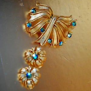 May include: A vintage gold-coloured brooch and matching earrings set. The brooch is bow-shaped with layered ribbons and teal-coloured rhinestones. The earrings are flower-shaped with similar teal rhinestones. The jewellery set is on a gold-coloured surface.