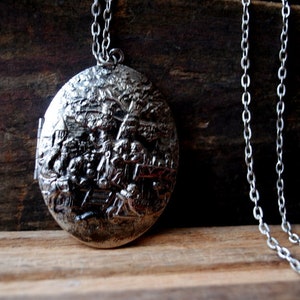 May include: Silver-toned oval locket pendant with a detailed relief scene, suspended from a delicate chain. The locket features a hinged design and intricate artwork, creating a vintage aesthetic. The chain is approximately 20 inches long.