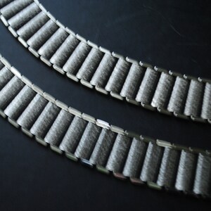 May include: Two gray watch bands with a textured, ribbed design. The bands are made of a soft, felt-like material and are connected by silver-colored metal links. The bands are arranged in a curved pattern against a dark background.