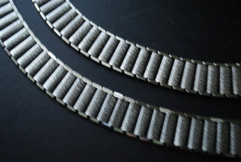 May include: Two gray watch bands with a textured, ribbed design. The bands are made of a soft, felt-like material and are connected by silver-colored metal links. The bands are arranged in a curved pattern against a dark background.