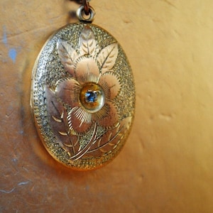 May include: A gold oval locket with a detailed floral design and a blue gemstone in the center.