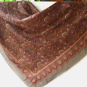 May include: A square scarf in a muted brown tone featuring a repeating floral and vine pattern in shades of deep red, orange, and cream. The design includes a distinct border of stylized flower motifs along the edges with a soft, textured fabric.