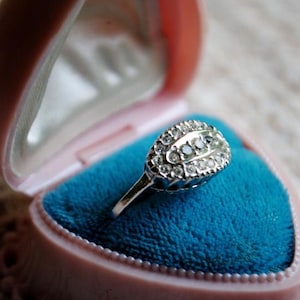 May include: A silver ring with a cluster of small, clear gemstones in an oval shape, set in a pink jewelry box with a blue velvet interior. The ring is displayed on a textured, off-white surface.