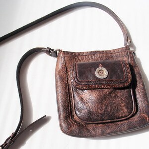 May include: A brown leather crossbody bag with a flap closure and a silver button. The bag has a distressed look and a small pocket on the front.