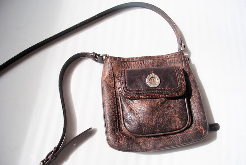 May include: A brown leather crossbody bag with a flap closure and a silver button. The bag has a distressed look and a small pocket on the front.