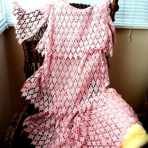 May include: A light pink, open-weave lace dress with bell sleeves and a tiered skirt. The dress is draped over a wicker chair with a fur throw. The intricate lace pattern is visible throughout the garment.