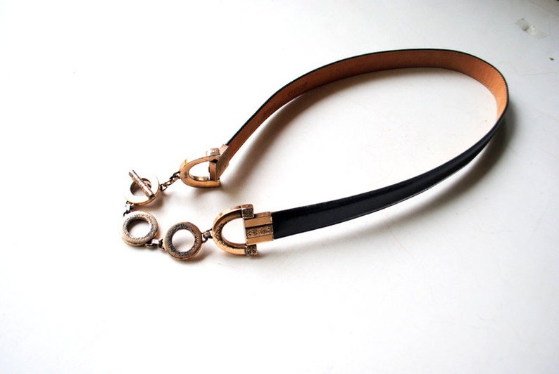 Peut inclure: Une ceinture en cuir noir avec une boucle dor&eacute;e et un d&eacute;tail de cha&icirc;ne. La boucle pr&eacute;sente un motif d&eacute;coratif.