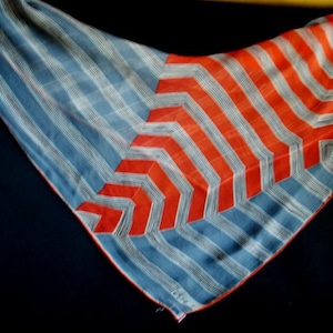 Op de afbeelding: Een vintage zijden sjaal met een opvallend geometrisch ontwerp. De sjaal heeft een blauw-wit gestreepte achtergrond met een vet oranje chevronpatroon. De sjaal heeft een driehoekige vorm.