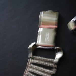 May include: Close-up of a silver-toned watch band clasp and a portion of the watch band. The clasp is rectangular with a hinged closure. The watch band is made of small, connected metal links. The background is a dark gray.