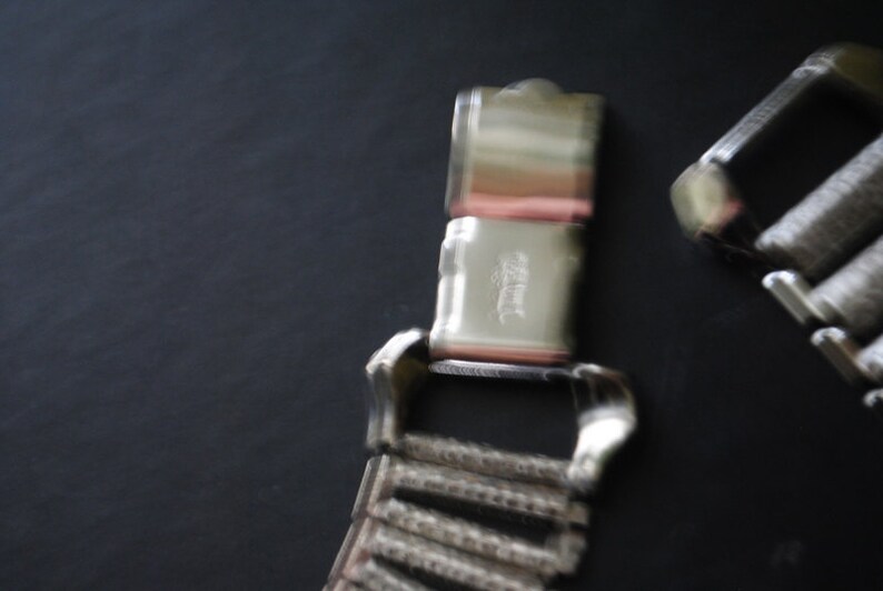 May include: Close-up of a silver-toned watch band clasp and a portion of the watch band. The clasp is rectangular with a hinged closure. The watch band is made of small, connected metal links. The background is a dark gray.