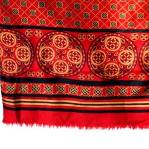 Op de afbeelding: Een rode textiel met een ingewikkeld patroon. Het ontwerp bevat herhaalde cirkelvormige medaillons in goud, rood en zwart. Het bovenste gedeelte heeft een ruitpatroon. De onderste rand is gerafeld.