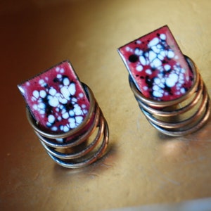 May include: A pair of earrings with a red and white enamel design. The earrings are made of copper and have a spiral shape.