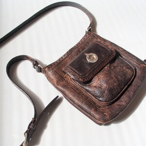 May include: A brown leather crossbody bag with a flap closure and a silver buckle. The bag has a distressed look and a small pocket on the front.