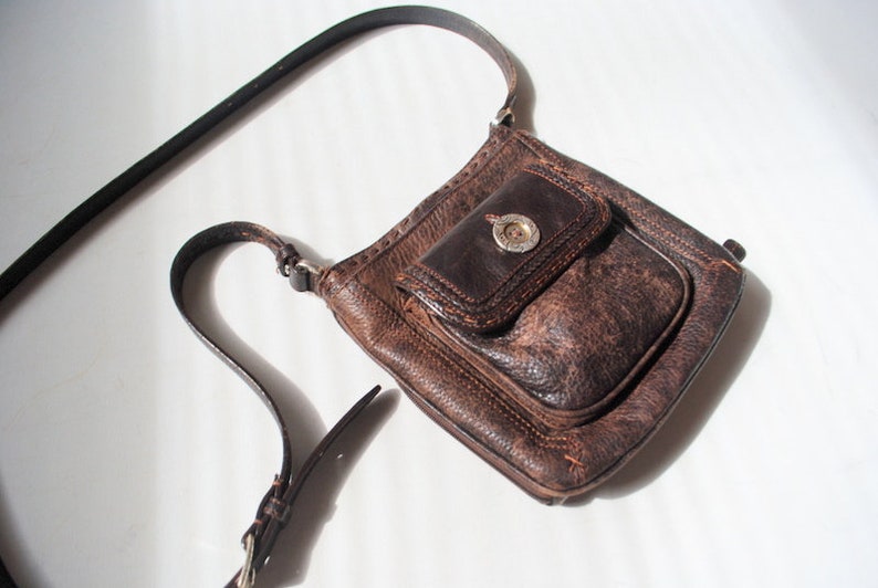 May include: A brown leather crossbody bag with a flap closure and a silver buckle. The bag has a distressed look and a small pocket on the front.