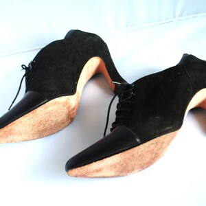 May include: A pair of black suede ankle boots with lace-up fronts and high heels. The heels are made of wood and have a natural finish.