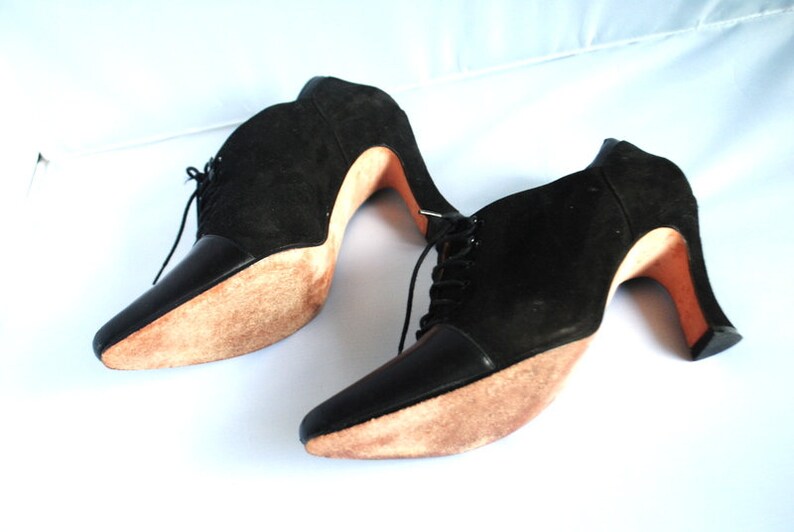 May include: A pair of black suede ankle boots with lace-up fronts and high heels. The heels are made of wood and have a natural finish.