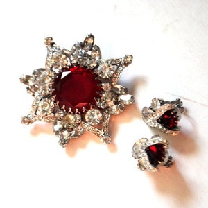 May include: A red rhinestone brooch and earrings set. The brooch is a starburst design with a large red stone in the center. The earrings are small and feature a red stone surrounded by rhinestones.