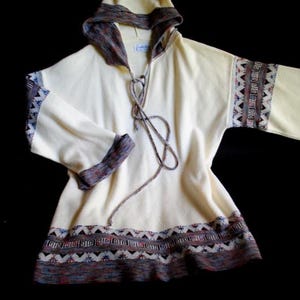 May include: Cream-colored hooded tunic with patterned trim on the sleeves, cuffs, and hem. The trim features geometric designs in shades of brown, blue, and red. The tunic has a drawstring neckline and long sleeves.
