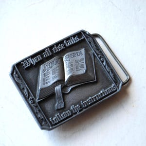May include: A rectangular, silver-colored belt buckle featuring an open book with the word "HOLY BIBLE" embossed on the pages. The buckle has the text "When all else fails... follow the instructions" around the edges.