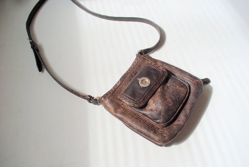 May include: A brown leather crossbody bag with a flap closure and a silver button. The bag has a small pocket on the front.