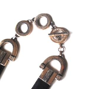 Peut inclure: Une ceinture en cuir noir avec une boucle dor&eacute;e. La boucle est orn&eacute;e et pr&eacute;sente un motif g&eacute;om&eacute;trique. La ceinture est ferm&eacute;e par un fermoir dor&eacute;.