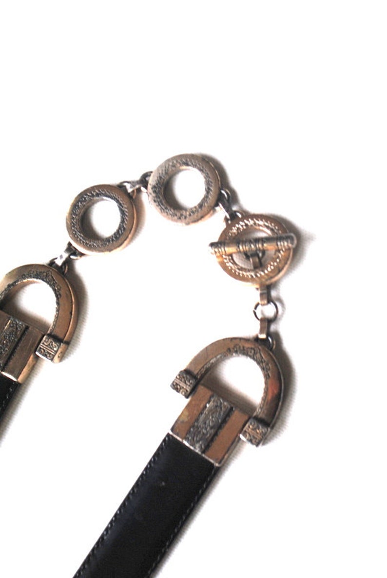 Peut inclure: Une ceinture en cuir noir avec une boucle dor&eacute;e. La boucle est orn&eacute;e et pr&eacute;sente un motif g&eacute;om&eacute;trique. La ceinture est ferm&eacute;e par un fermoir dor&eacute;.