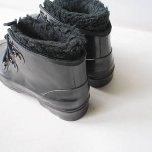 May include: A pair of black rubber boots with a faux fur lining. The boots have a lace-up design with metal eyelets and a rounded toe. The boots are on a white surface.