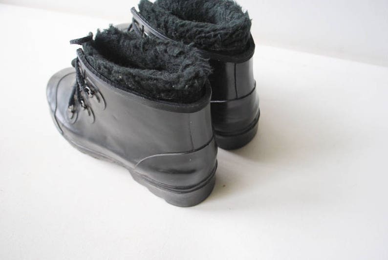 May include: A pair of black rubber boots with a faux fur lining. The boots have a lace-up design with metal eyelets and a rounded toe. The boots are on a white surface.