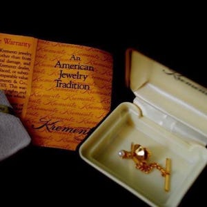 May include: A gold tie tack with a single pearl and a diamond-shaped design in a white box. The box has the words "An American Jewelry Tradition" and "Krementz" printed on it.