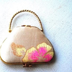 May include: A small, vintage beige handbag with a gold-colored twisted handle and trim. The bag features an embroidered floral design in shades of pink and gold. The clasp is gold-colored.