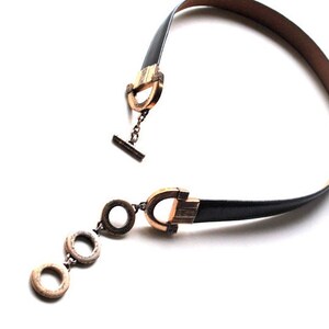 Peut inclure: Un collier ras de cou en cuir noir avec des fermoirs dor&eacute;s et un pendentif avec trois anneaux circulaires.