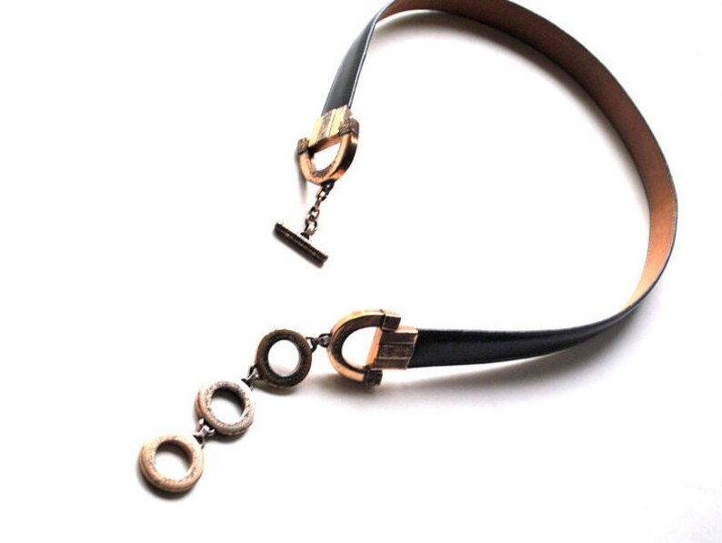 Peut inclure: Un collier ras de cou en cuir noir avec des fermoirs dor&eacute;s et un pendentif avec trois anneaux circulaires.