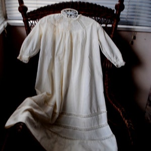 Pode incluir: Camisola de noite vintage creme, com mangas compridas e delicadas rendas. A camisola de noite é exibida numa cadeira de vime, mostrando o seu design fluido e estilo clássico. O tecido parece macio e leve.