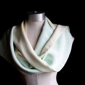 May include: A light green and cream coloured scarf draped over a mannequin. The scarf is made of a soft, flowing fabric.
