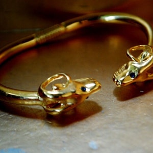 May include: A gold bracelet with two ram heads on the ends. The ram heads have black gemstones for eyes.
