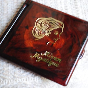 May include: A vintage compact mirror with a tortoiseshell pattern. The mirror features a gold-colored profile of a woman and the words "Mirror Mystique". The compact is square and has a dark brown border.