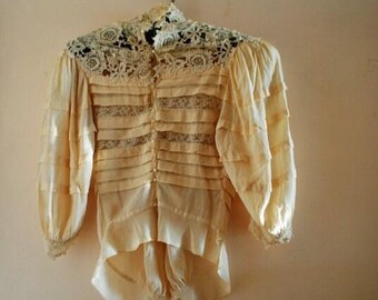 May include: A pale yellow blouse featuring a high lace collar, long puffed sleeves, and a front button closure. The bodice is constructed with alternating horizontal bands of solid fabric and intricate lace inserts, finished with a ruffled hem.