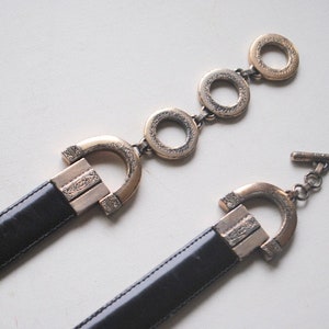 Peut inclure: Une ceinture en cuir noir avec deux boucles orn&eacute;es dor&eacute;es. L'une des boucles est un grand ovale avec un motif d&eacute;coratif et l'autre est une boucle plus petite et rectangulaire avec un motif similaire.