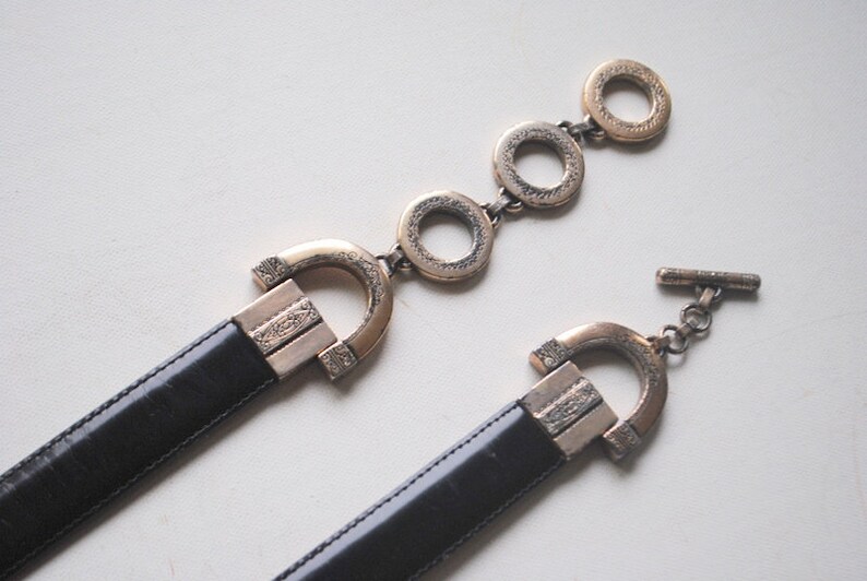 Peut inclure: Une ceinture en cuir noir avec deux boucles orn&eacute;es dor&eacute;es. L'une des boucles est un grand ovale avec un motif d&eacute;coratif et l'autre est une boucle plus petite et rectangulaire avec un motif similaire.