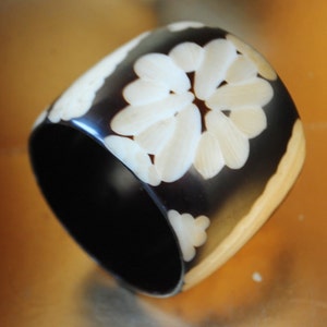 May include: A black and white bangle bracelet with a carved floral design. The bracelet is made of horn and has a polished finish.