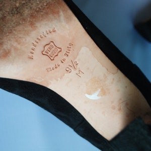 May include: Close-up of the inside of a black suede shoe. The insole is tan leather with the words "handcrafted" and "Vero Cuoio" stamped in brown. The size is marked as "9 1/2 M" in brown.