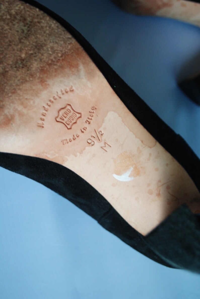 May include: Close-up of the inside of a black suede shoe. The insole is tan leather with the words "handcrafted" and "Vero Cuoio" stamped in brown. The size is marked as "9 1/2 M" in brown.