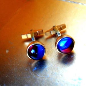 May include: A pair of cufflinks with a gold-coloured metal setting and a round, cabochon-style stone. The stones display a vibrant mix of blue and red hues, creating a striking visual contrast. The cufflinks are designed for formal wear.