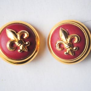 Op de afbeelding: Een paar gouden clip-oorbellen met rood emaille en een gouden fleur-de-lis-ontwerp.