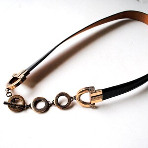 Peut inclure: Une ceinture en cuir noir avec une boucle dor&eacute;e et des maillons de cha&icirc;ne d&eacute;coratifs. La boucle pr&eacute;sente un design unique avec une forme circulaire et des d&eacute;tails complexes.