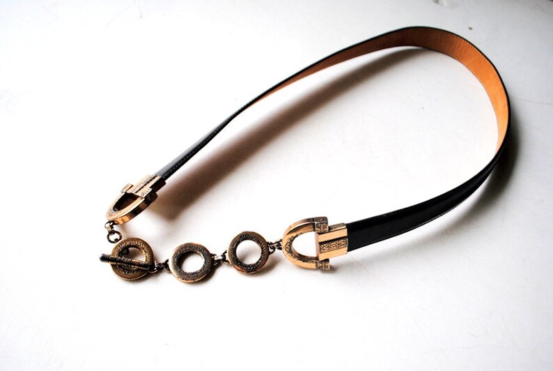 Peut inclure: Une ceinture en cuir noir avec une boucle dor&eacute;e et des maillons de cha&icirc;ne d&eacute;coratifs. La boucle pr&eacute;sente un design unique avec une forme circulaire et des d&eacute;tails complexes.