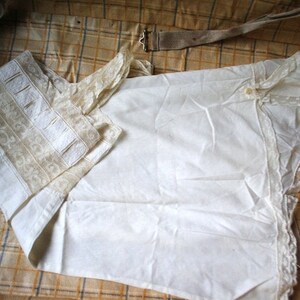 May include: A white cotton garment with lace trim and a beige embroidered collar. The garment is folded and lying on a plaid fabric surface.