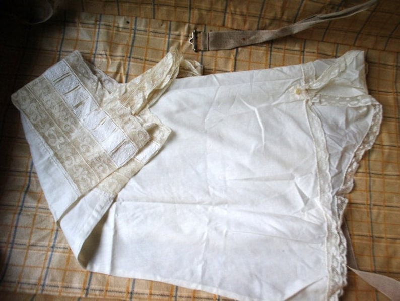 May include: A white cotton garment with lace trim and a beige embroidered collar. The garment is folded and lying on a plaid fabric surface.